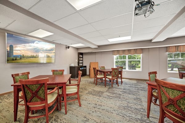 Spacious common area with wooden tables and chairs, featuring a large wall-mounted screen.