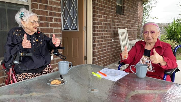 Two senior women sitting outside, smiling and giving thumbs up.