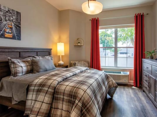 A well-decorated bedroom featuring a plaid blanket, lamp, and window with red curtains.