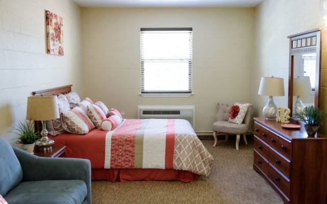 A well-decorated bedroom featuring a bed with colorful pillows, a dresser, and a cozy chair in a senior living facility.