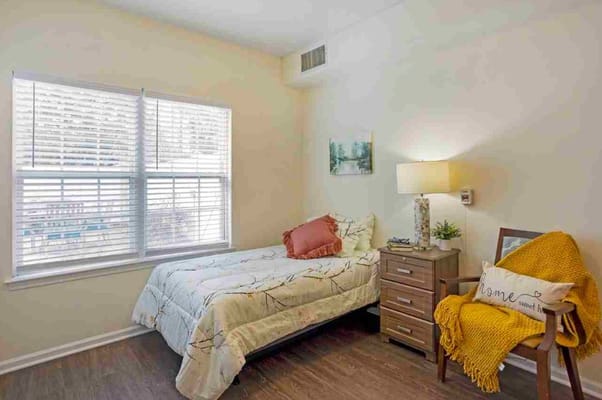 A cozy bedroom with a bed, lamp, and chair in Floral Creek Alzheimer’s Special Care Center.