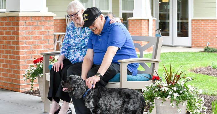Two senior residents sitting on a bench, petting a dog in a garden area.