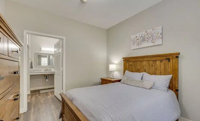 A neatly arranged bedroom featuring a wooden bed and dresser with a bathroom in the background.