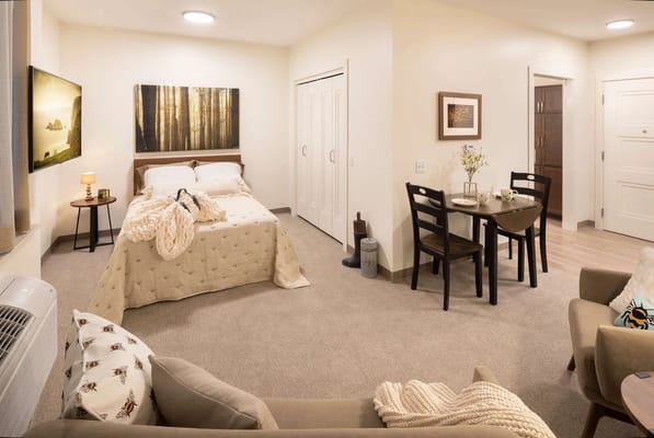 A well-furnished bedroom with a bed, dining table, and easy chair in TLC Assisted Living.