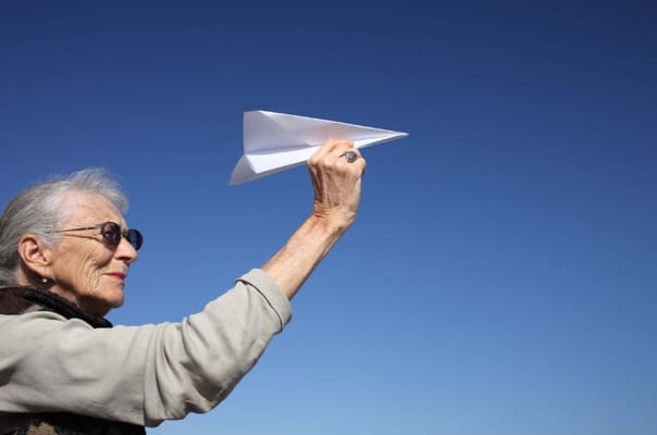 An elderly woman holding a paper airplane against a clear blue sky.