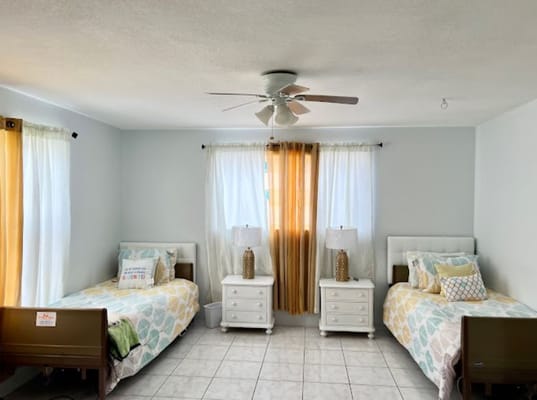 A bright bedroom with two beds, lamps, and large windows.
