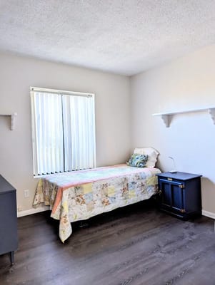 A cozy bedroom featuring a single bed, bedside table, and window with blinds.