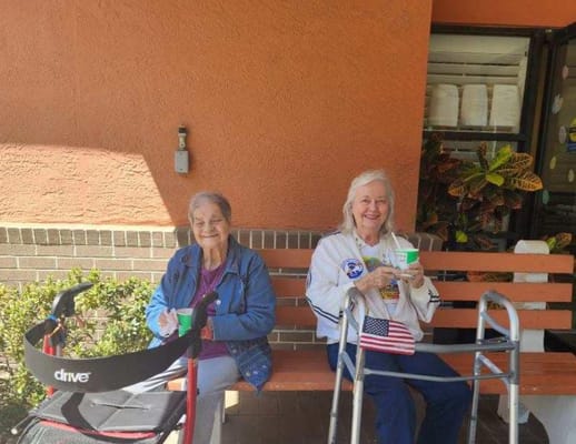 Two seniors sitting on a bench with drinks outside the facility.