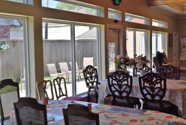 Bright dining area with wooden tables and chairs, flowers, and large windows.