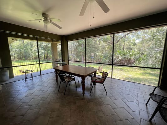 A spacious screened porch with a wooden table and chairs, surrounded by greenery.