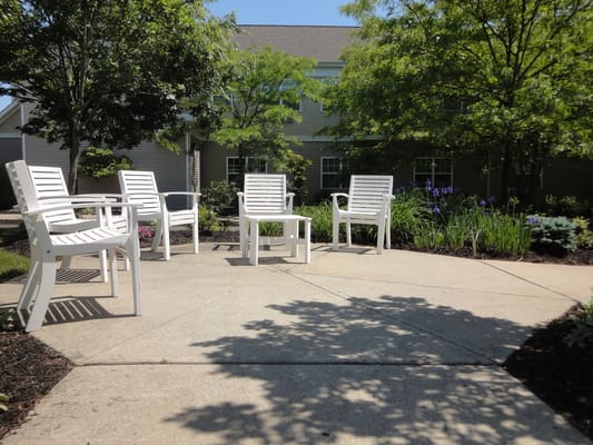 White patio chairs arranged around a concrete area with greenery in the background