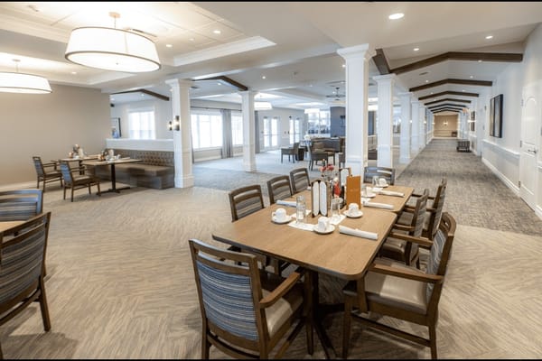 A dining area in Vitalia Senior Residences featuring tables and chairs, with a view of the hallway.