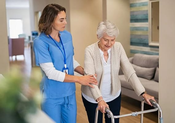 A caregiver assists a senior woman using a walker in a hallway.