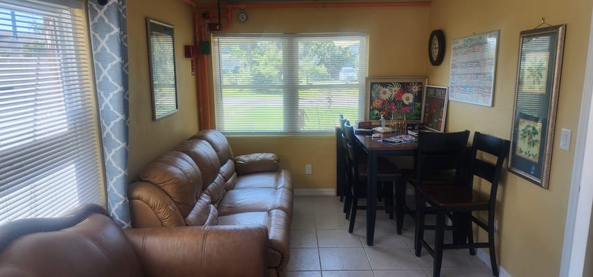 A living room with a brown sofa and dining area featuring a window and decorative artwork.
