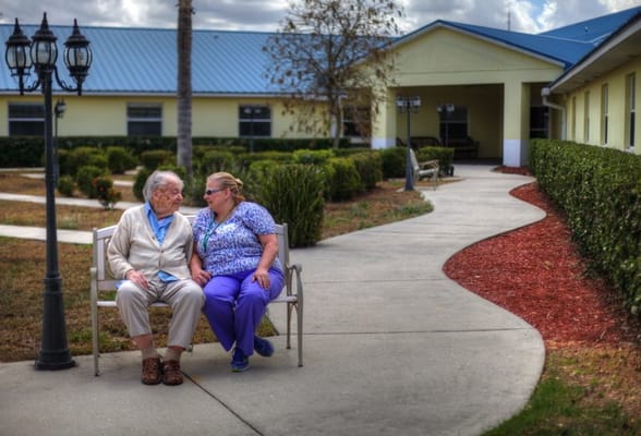 An elderly man and a caregiver seated on a bench, engaged in conversation in a landscaped area.