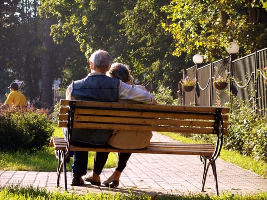 Elderly couple sitting close together on a park bench in a sunny area.