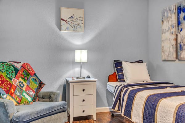 A cozy room featuring a bed, nightstand, and armchair with a colorful blanket.