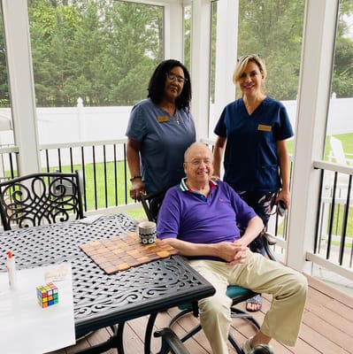 Two caregivers smiling beside a seated senior man at a table on a porch