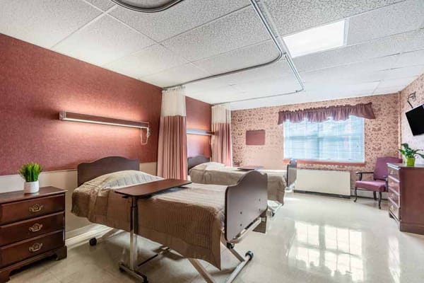 A shared bedroom featuring two adjustable hospital beds, a nightstand, and natural light from a window.