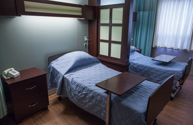 A shared bedroom with two hospital beds and wooden furniture in a senior living facility.