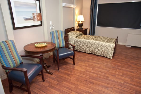Interior of a senior living room with a bed, two chairs, and a table.