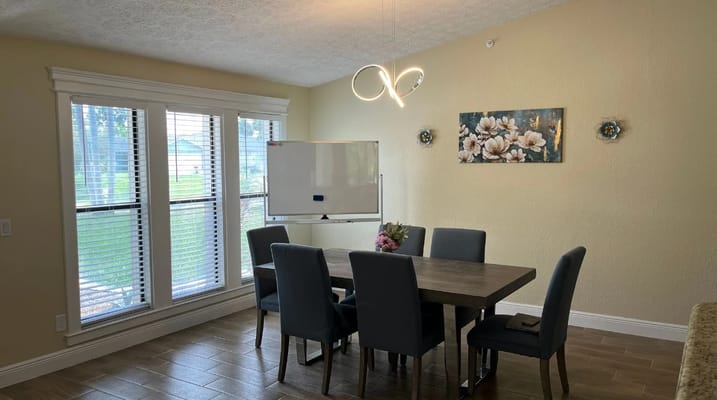 A well-lit dining room with a table and six chairs, featuring a whiteboard and floral wall art.