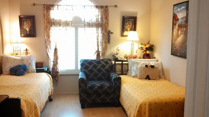 A cozy bedroom with two beds and a chair, decorated with floral curtains and artwork.