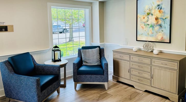 A cozy living room area with two blue chairs, a side table, and a floral painting.