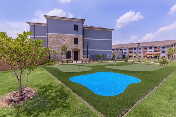 A putting green with artificial turf and a small blue pond in The Retreat Senior Living garden.