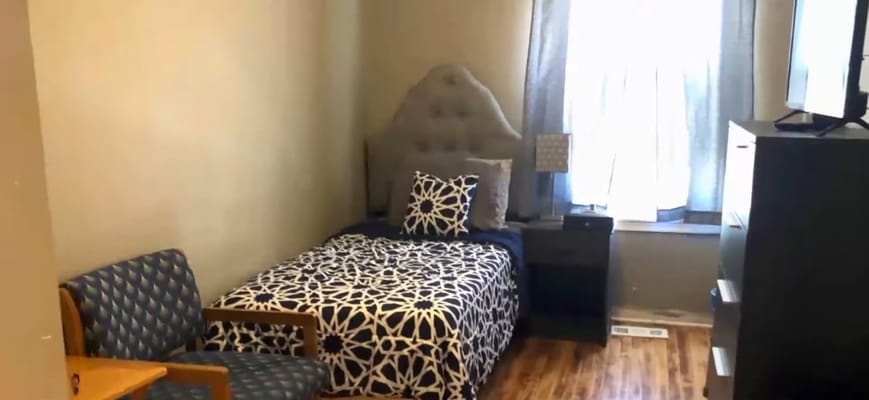 A modestly decorated resident room featuring a single bed, a bedside table, and a chair.