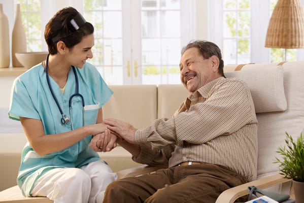 A caregiver smiles at a senior resident while holding hands in a cozy living area.