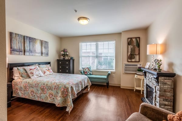 A cozy bedroom featuring a floral bedspread, dresser, and natural light from a window.