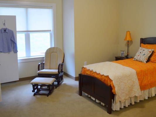 A cozy bedroom featuring a bed with an orange coverlet, a rocking chair, and a lamp.