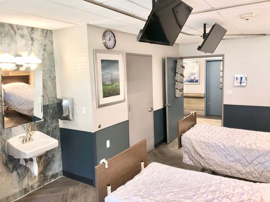 Patient bedroom with two beds, wall-mounted TVs, and a sink.