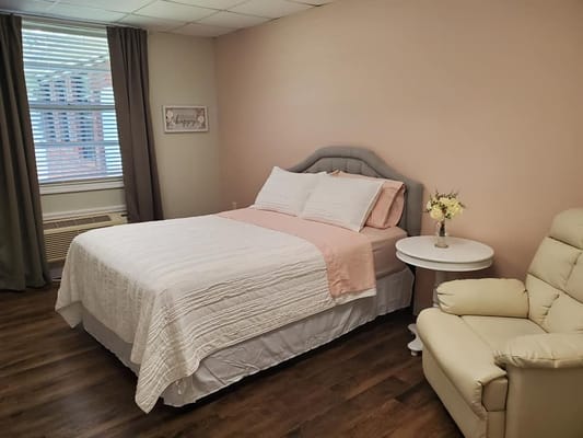 A cozy bedroom featuring a bed with cream and pink bedding, a side table with flowers, and a recliner.