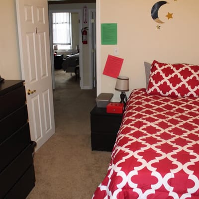A neatly arranged bedroom with a red patterned bedspread and a view into another room.