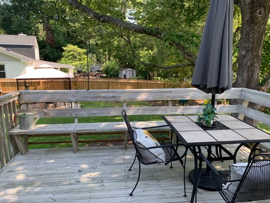 Wooden deck with a table and chairs, surrounded by greenery.