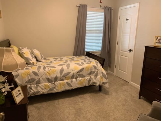 A well-furnished bedroom with floral bedding and a window covered by light curtains.