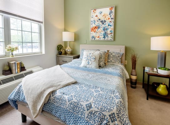 A cozy bedroom featuring a bed with blue and white patterned bedding, a lamp, and artwork on the wall.