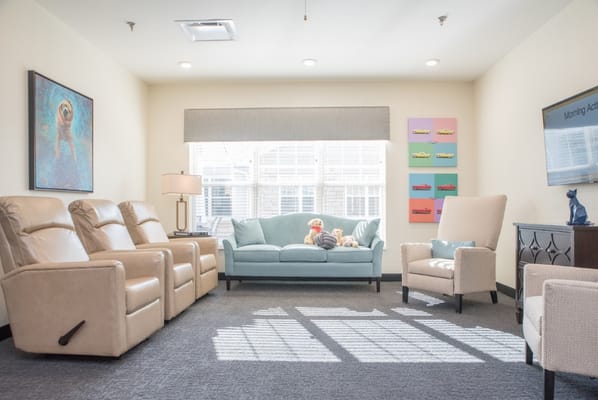 A cozy living room with a blue sofa, recliners, and bright sunlight streaming through the windows.