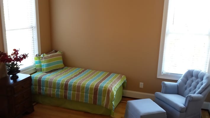 A cozy bedroom featuring a striped bedspread, a blue chair, and a window with natural light.