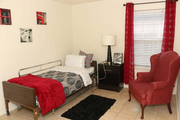 A cozy bedroom featuring a hospital bed with red throw blanket, a nightstand, lamp, and red curtains.