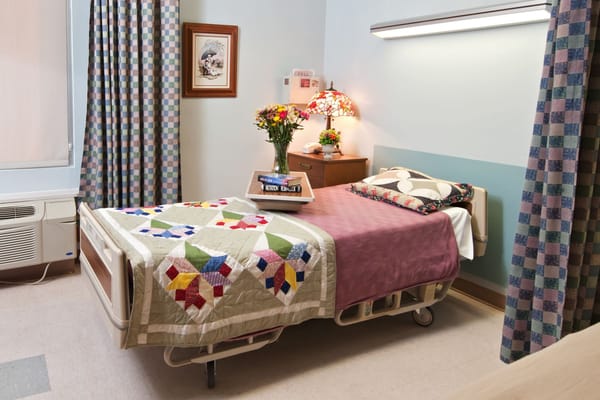A spacious patient bedroom with a quilted bedspread, vibrant flowers, and decorative curtains.