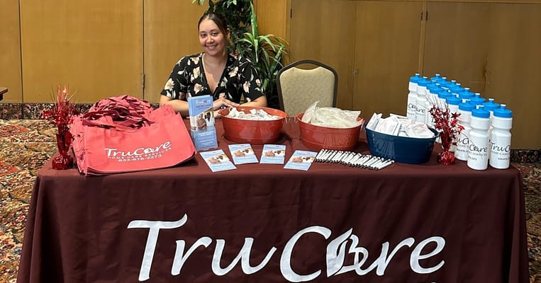 Staff member at TruCare booth with promotional materials and giveaways.