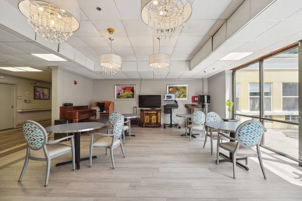 A bright common area featuring tables, chairs, and a piano.