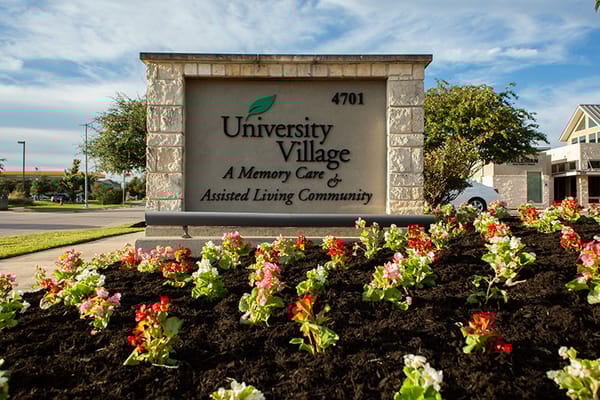 Sign for University Village Memory Care and Assisted Living Community with flowers in the foreground.