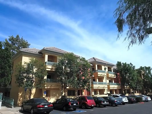Three-story building with balconies surrounded by trees and parked cars