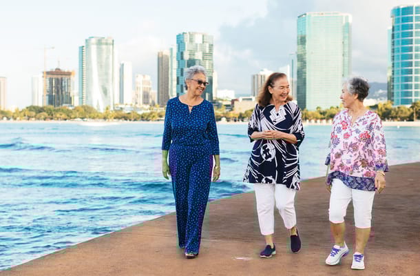 Three senior women walking by the water with a city skyline.