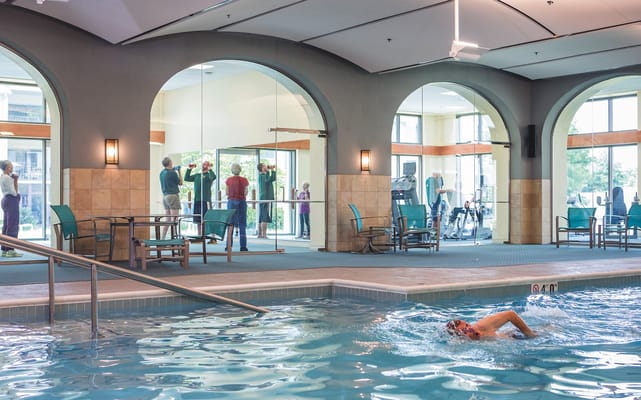 Indoor swimming pool with a swimmer in the foreground and fitness center visible through glass walls.