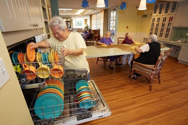 A resident loading colorful dishes into a dishwasher while others sit at a table.